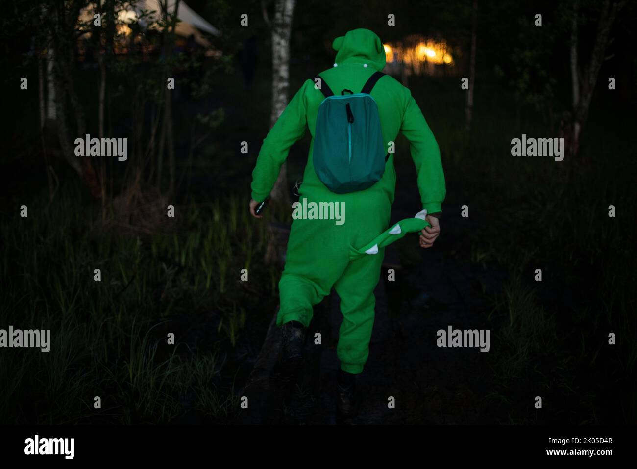 Man in green suit walks through woods. Guy in fun crocodile costume. Man at music festival in nature. Stock Photo