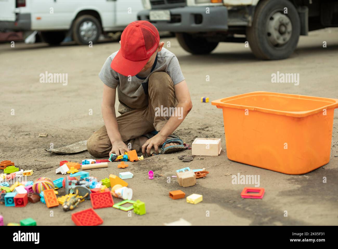 Child collects toys in drawer. Boy scattered toys on road. Preschooler boy on street. Game in summer. Street child. Stock Photo