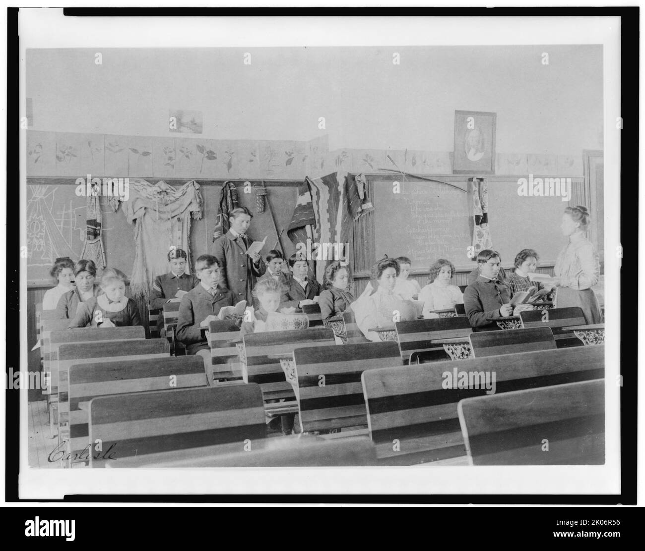 Carlisle, between 1901 and 1903. Classroom at the Indian Industrial School, Carlisle, Pennsylvania, showing teacher observing students reading. (Boarding school for Native American students, founded in 1879 under US governmental authority. Male and female students in class. Note items of traditional clothing attached to wall, and drawing of tipi on blackboard). Stock Photo