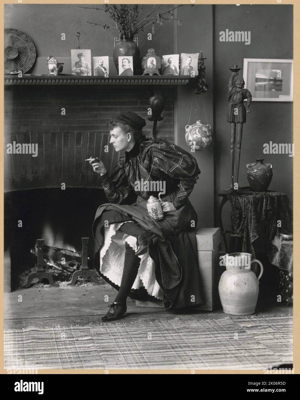 Frances Benjamin Johnston, full-length portrait, seated in front of fireplace, facing left, holding cigarette in one hand and a beer stein in the other, in her Washington, D.C. studio, 1896. In this self-portrait the photographer Frances Benjamin Johnston poses as an independent 'new woman.' On the mantlepiece are six portraits she took of men (from left to right): poet Bliss Carman; A. N. Brown, likely the librarian at the U.S. Naval Academy; Henry Guston Rogers, likely the inventor and playwright Henry Gustave Rogers; architect James Rush Marshall; Smithsonian librarian Frank Phister; and L. Stock Photo