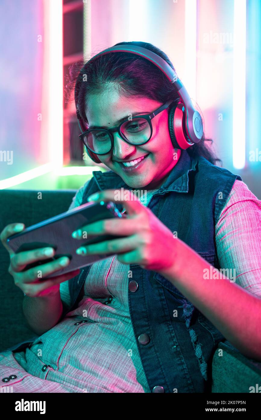 vertical shot of excited girl playing video game on mobile phone with wireless headset while sitting on sofa at home - concept of relaxation, hobby Stock Photo