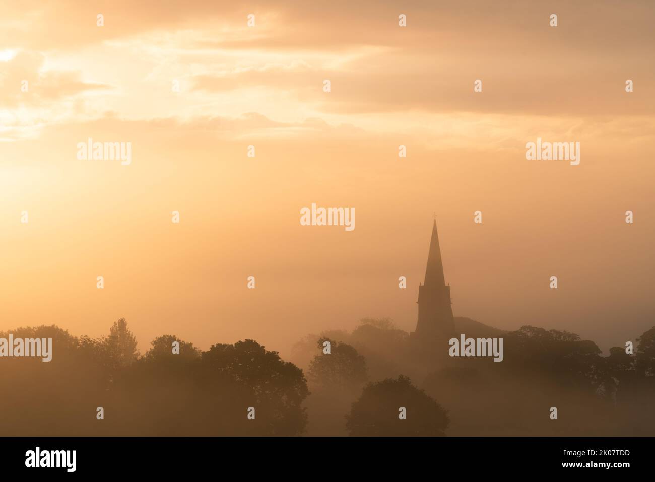 The steeple of St Barnabas Church in Weeton is silhouetted at sunrise on a foggy summer morning. Stock Photo