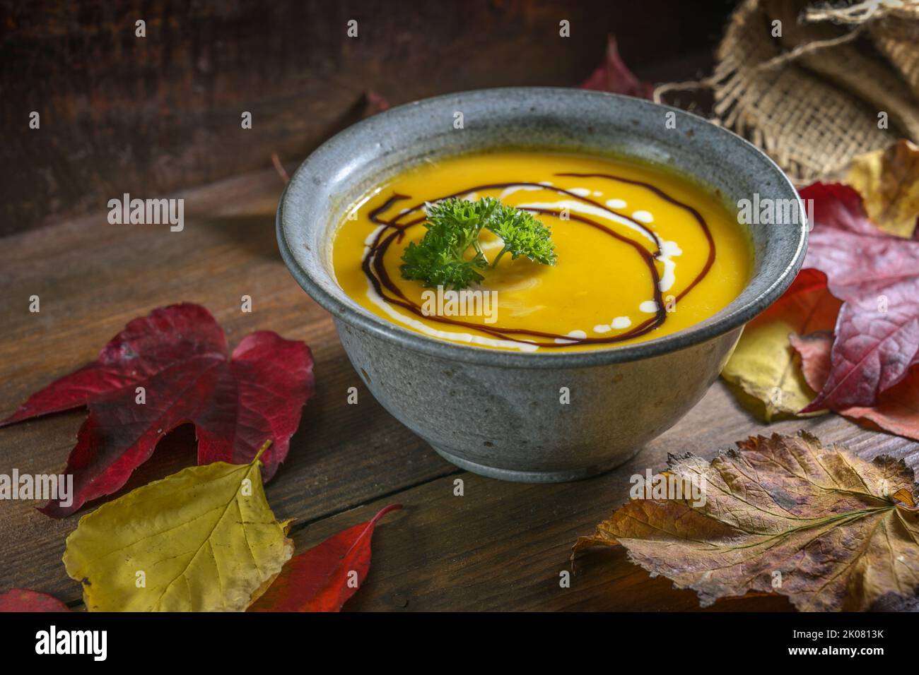 Autumn soup from red kuri squash with parsley garnish in a rustic bowl on a dark wooden table with colorful leaves for Thanksgiving or Halloween, copy Stock Photo
