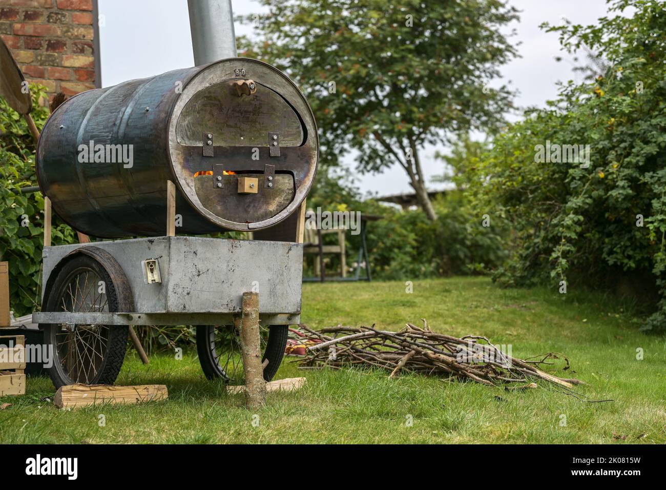 Homemade mobile pizza oven from an old metal barrel on a recycled chassis in a garden, fire in the combustion chamber and firewood on the lawn, copy s Stock Photo