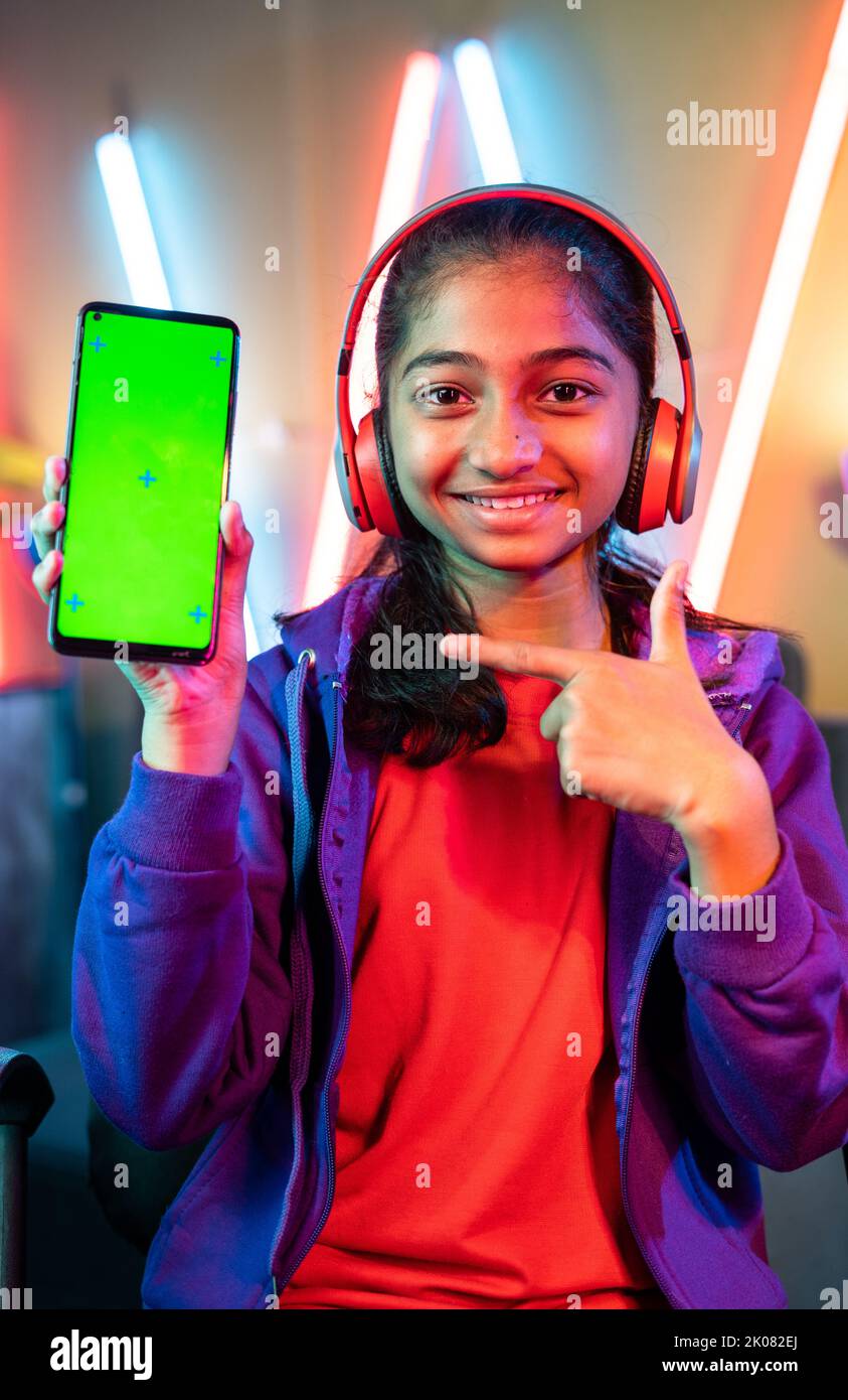 Vertical shot of Happy teenager girl kid with headset showing green screen mobile phone by pointing finger by looking camera at home - concept of app Stock Photo