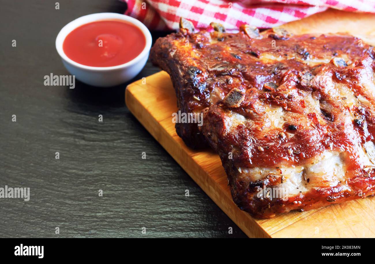 grilled pork ribs on a wooden cutting board with tomato ketchup on black slate surface Stock Photo