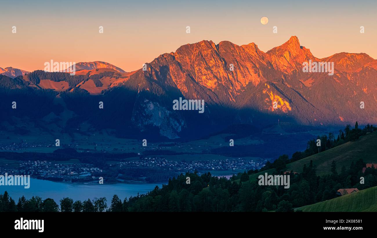 A beautiful summer morning during sunrise and the full moon slowly descending behind the mountains. The climatic health resort of Heiligenschwendi is Stock Photo