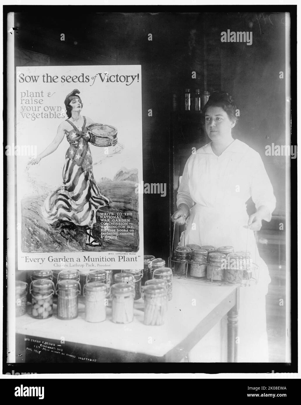 Mrs. Frank P. Brown, Cincinnati, Ohio. 1st prize winner of canned vegetables, Aug. 191[?]. First World War: woman with preseved food, and poster with personification of the USA, encouraging people to grown their own vegetables: 'Sow the seeds of Victory! plant and raise your own vegetables; write to the National War Garden Commission, Washington, D.C. for free books on gardening, canning and drying. Every Garden a Munition Plant - Charles Lathrop Pack, President'. Stock Photo