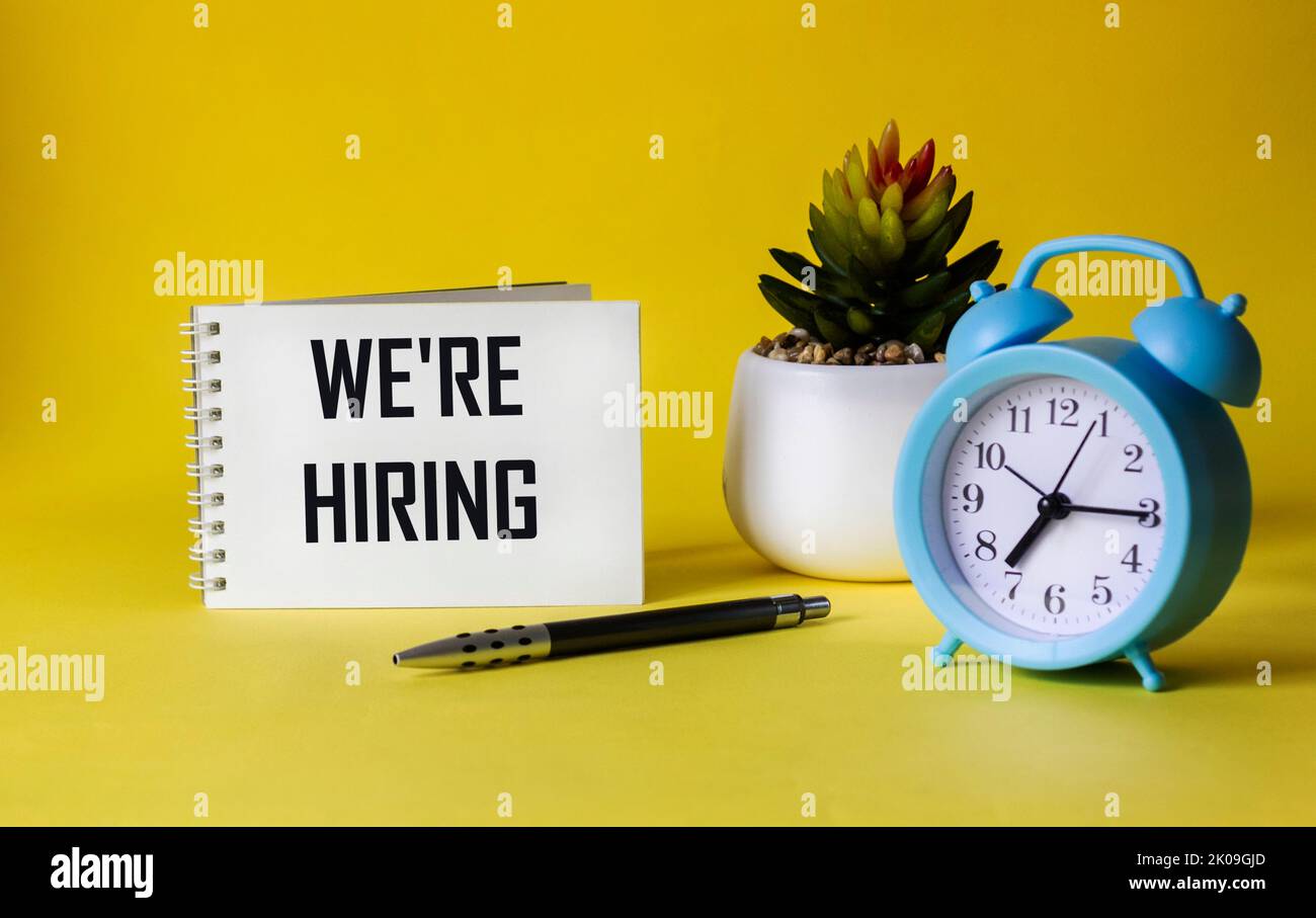 Notepad on a yellow background with the text We are hiring, next to a pen, a clock and a cactus Stock Photo
