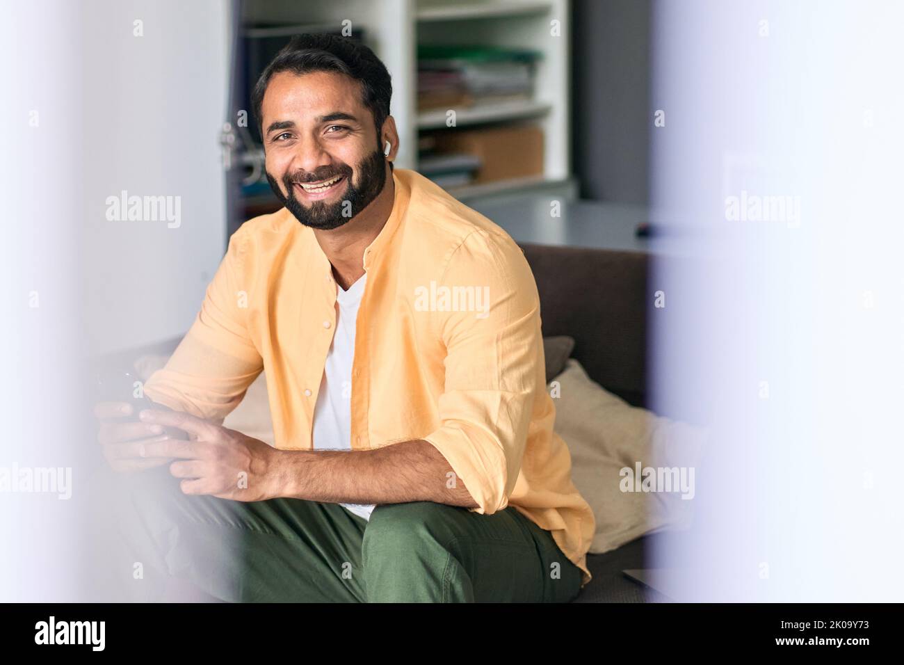 Happy indian man sitting at home on couch using phone listening music ...