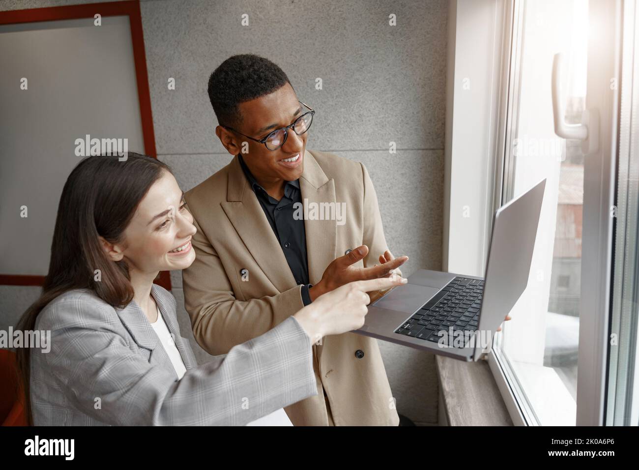 Two smiling business people working with laptop together standing near window Stock Photo