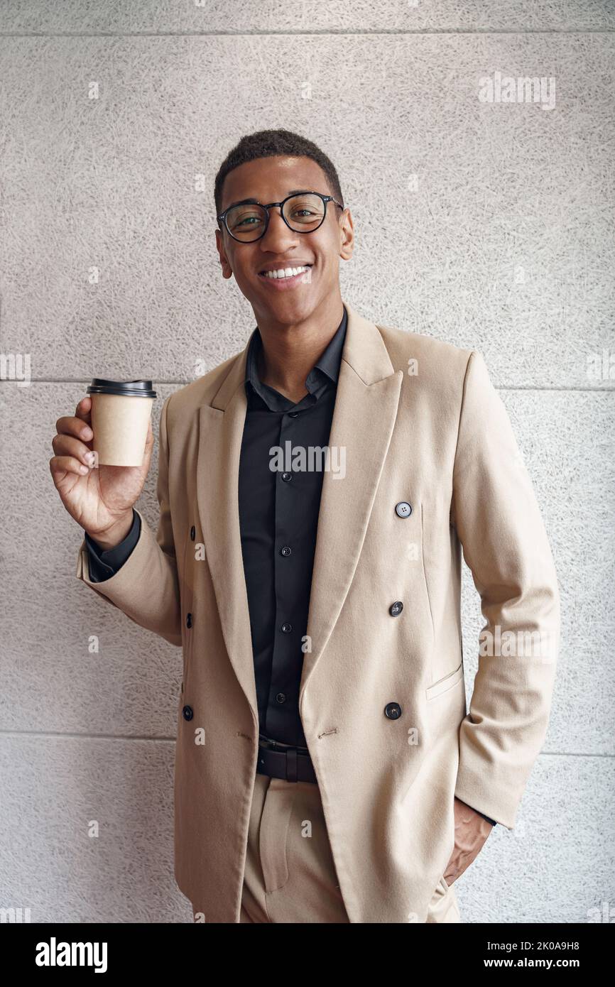 Handsome smiling african businessman with coffee standing near wall in modern coworking Stock Photo