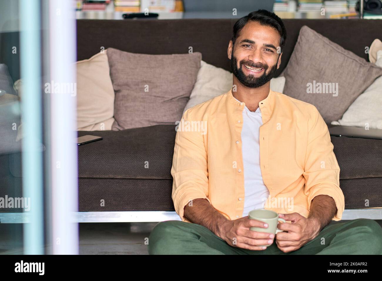 Happy indian man looking at camera drinking coffee sitting on sofa at