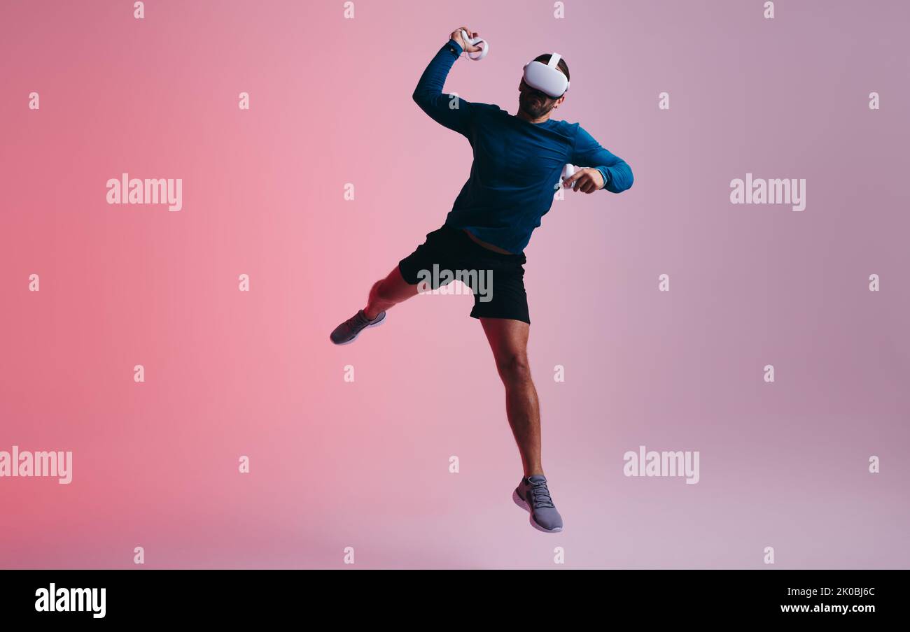 Energetic young man throwing a virtual ball using gaming controllers. Active young man jumping mid air while wearing virtual reality goggles. Young ma Stock Photo