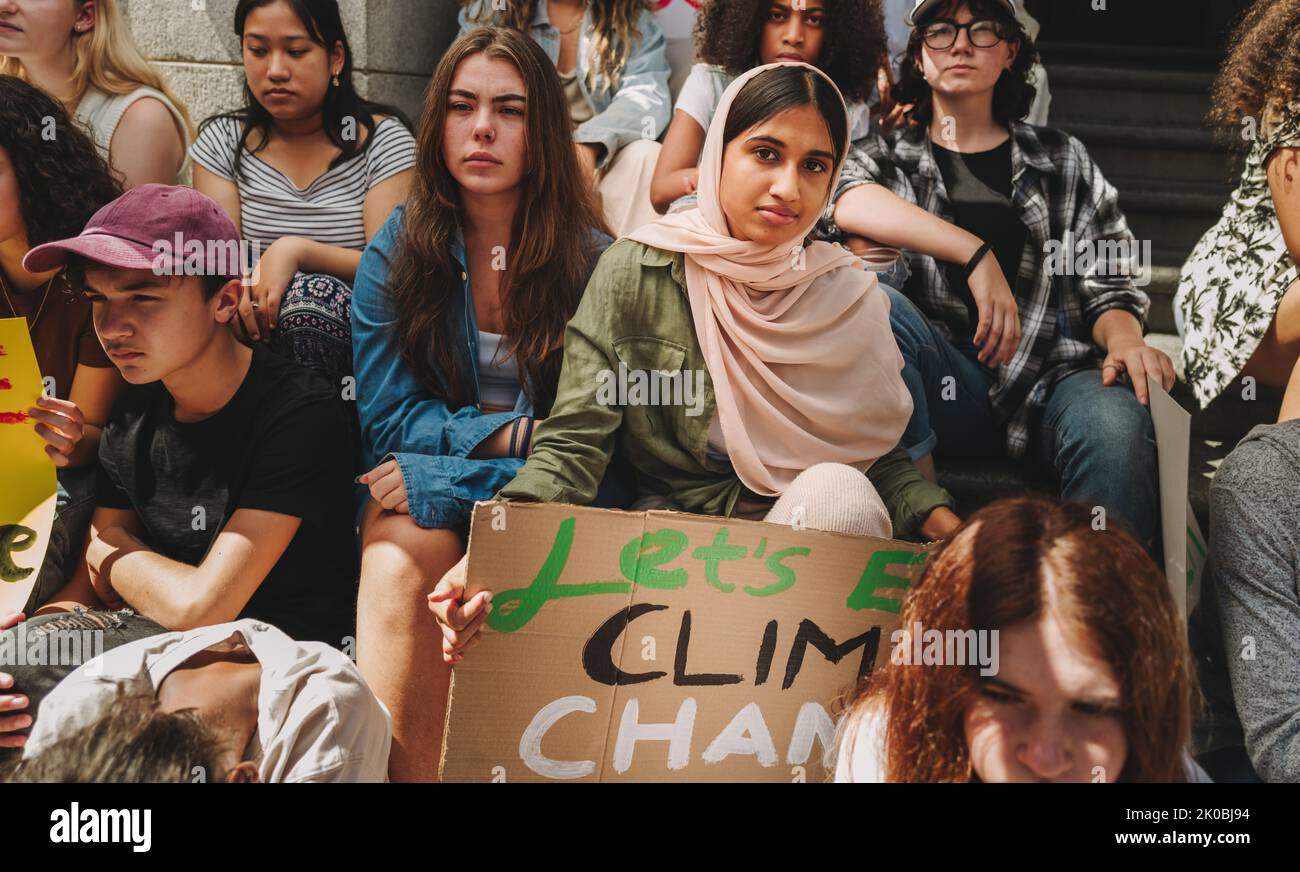 Generation Z climate activism. Young Muslim activist holding a poster while sitting with a group of demonstrators outside a building. Young people pro Stock Photo