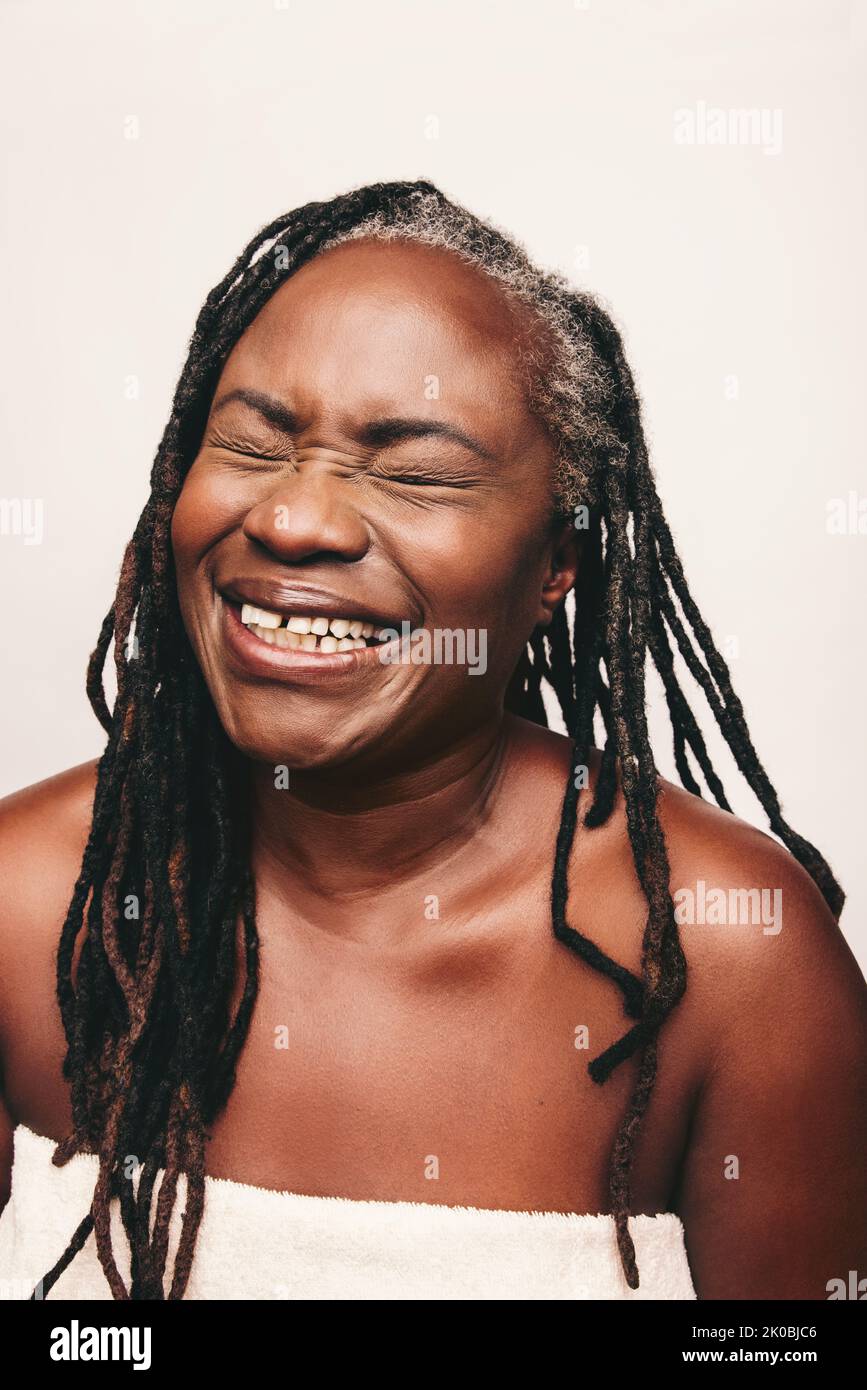 Joyful mature woman laughing cheerfully while wrapped in a bath towel. Refreshed woman with dreadlocks standing against a white background. Happy midd Stock Photo