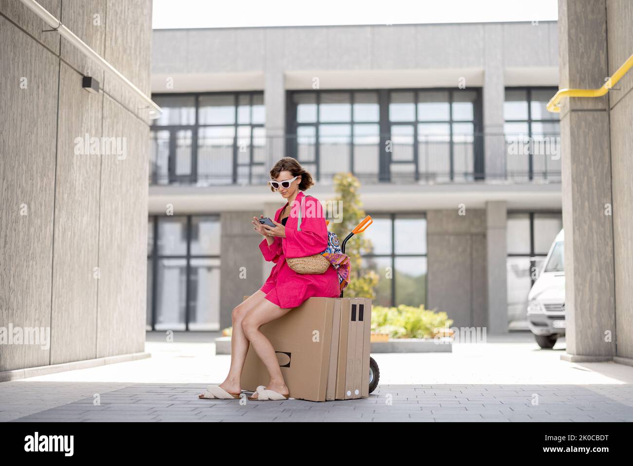 Woman using phone while sitting on push cart with furniture in boxes, delivering them home. Process of self improvement of apartment, buying furniture Stock Photo