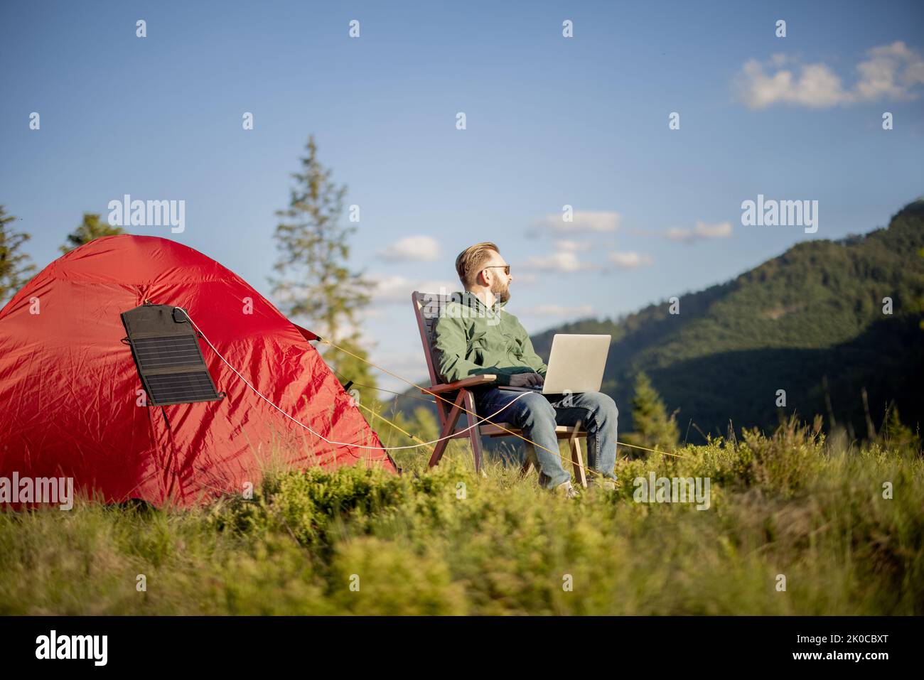Man works on laptop while traveling with tent in the mountains. Charging computer with portable solar panel hanging on tent. Concept of remote work at Stock Photo