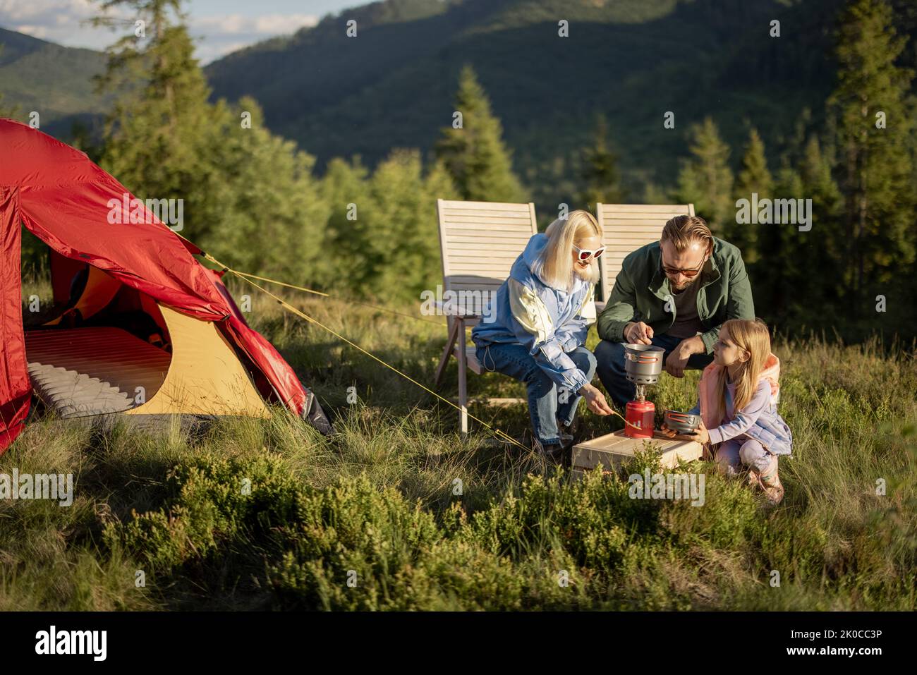 Young caucasian couple with little girl sit relaxed on chairs while traveling with tent in the mountains. Concept of happy family vacation on nature Stock Photo