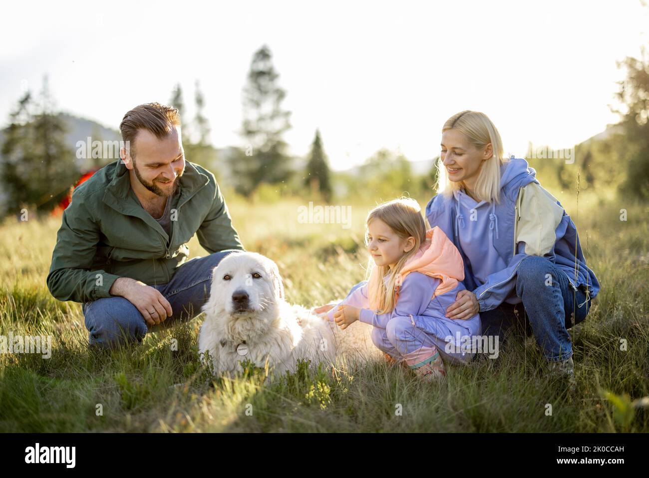 Portrait of a lovely couple with little girl and dog sit together while travel in the mountains. Young caucasian family spend summer vacation on natur Stock Photo