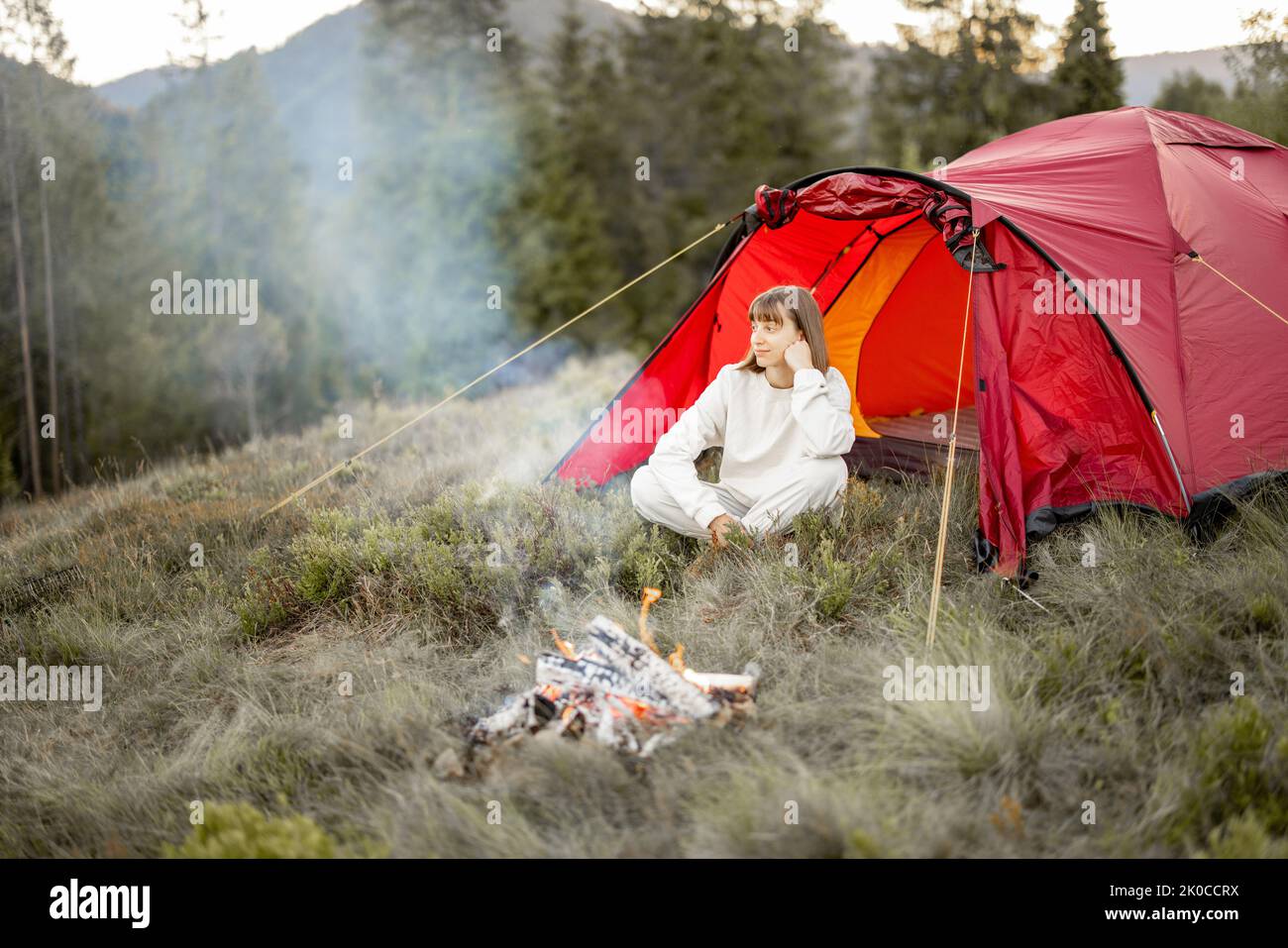 Young woman enjoys nature while sitting at campfire near camping tent in the mountains. Concept of escaping to nature Stock Photo