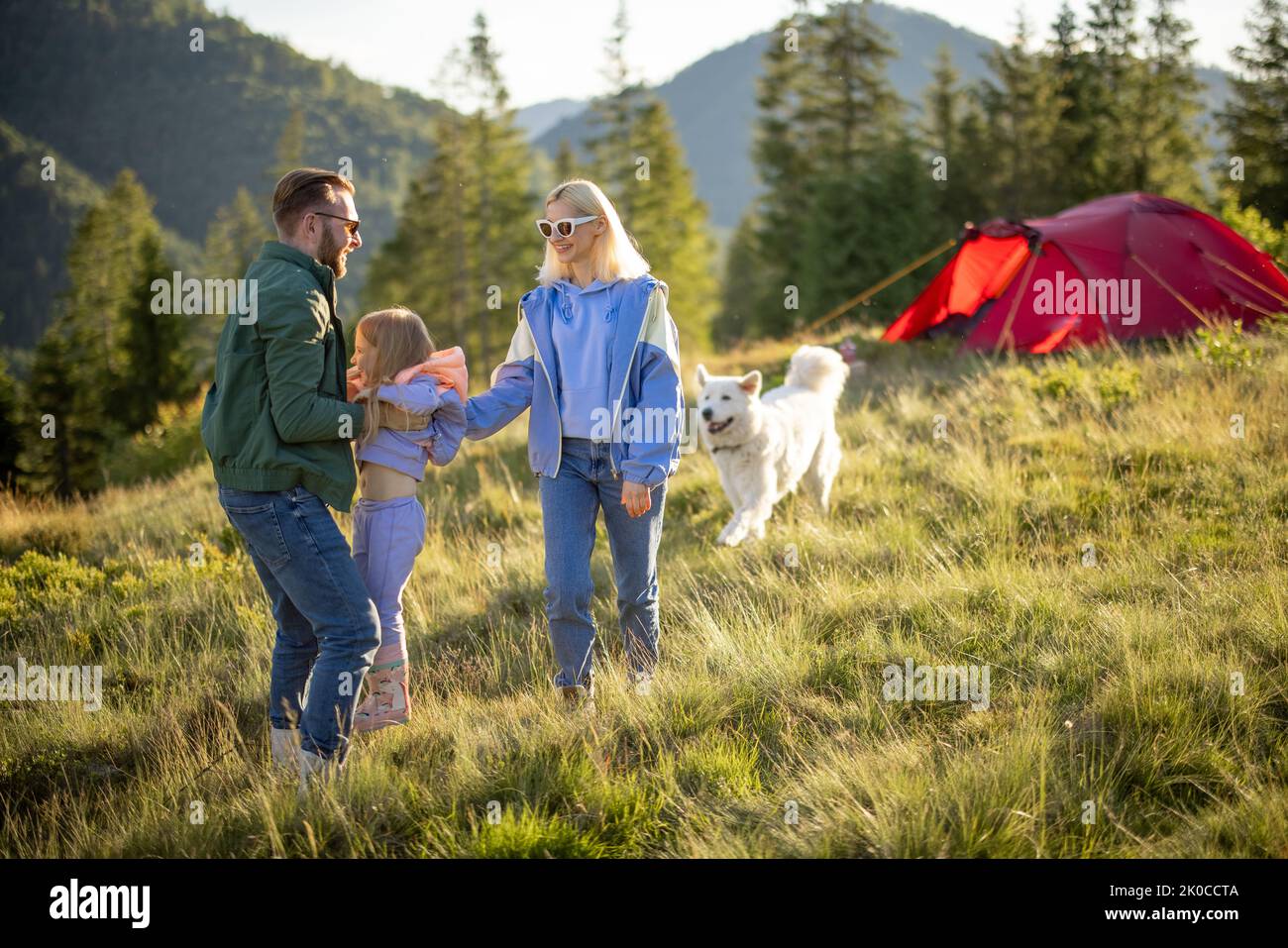 Young caucasian couple with little girl and dog have fun while travel with tent in the mountains. Happy family summer vacation at campsite. Father tos Stock Photo