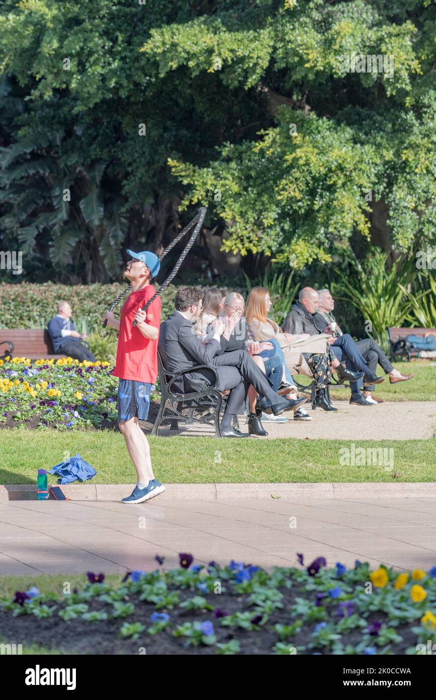 While people sit and eat their lunch in the sun, a man is skipping using a skipping rope in Hyde Park, Sydney, Australia Stock Photo