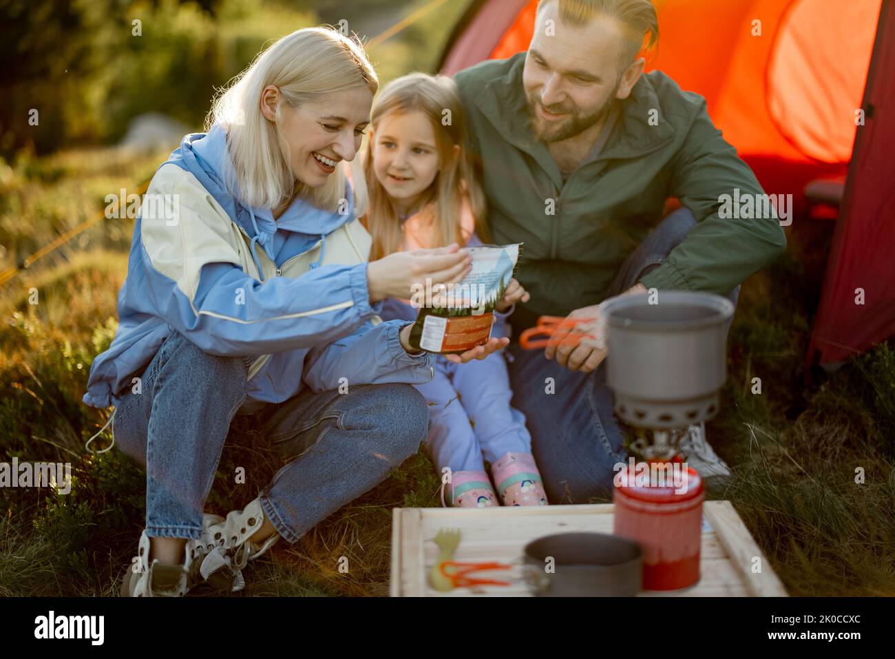 Young couple with little girl have picnic, cooking dried-food for hiking in package at campsite. Concept of sublimation food for travel and family bon Stock Photo