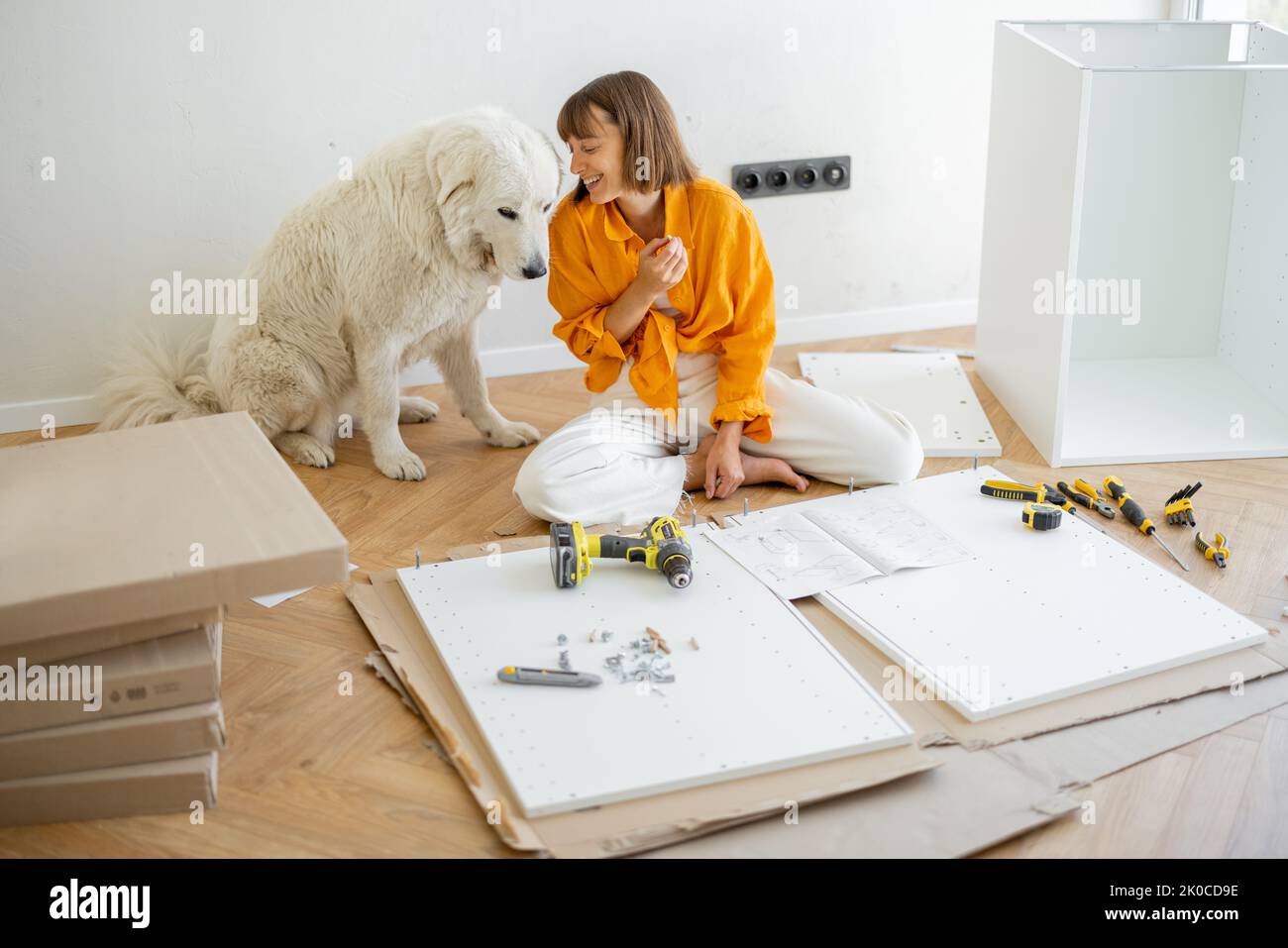 Young woman assembles furniture by herself, sitting and having fun with her cute dog at new apartment. DIY and house improvement concept Stock Photo