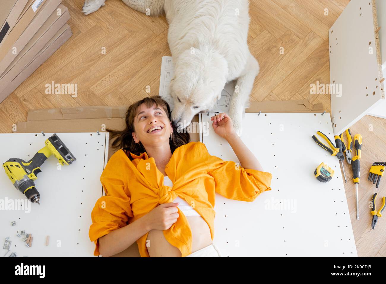 Young happy woman lies on floor with her cute dog, resting while making repairing at new apartment, top view. Young woman assembling furniture by hers Stock Photo