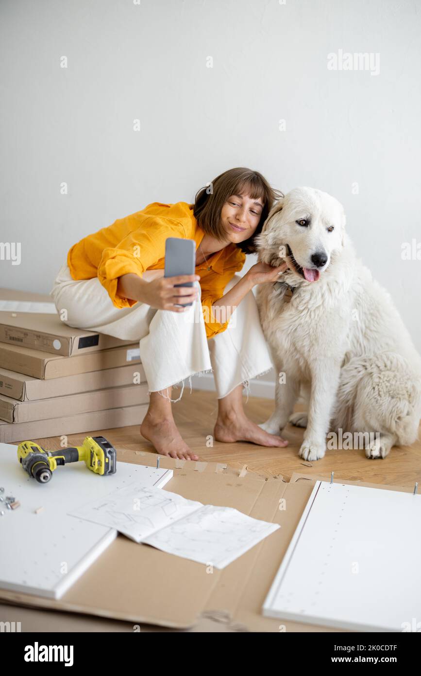 Young woman makes selfie photo on phone with her cute dog, have fun while making repairing at new apartment. Happy improvement and house repairing con Stock Photo