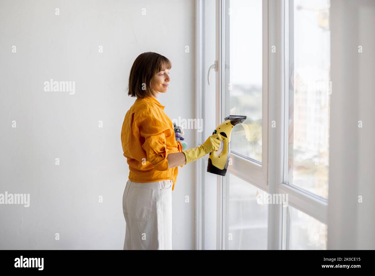 Young housewife or a cleaning company employee washes window with special tool in apartment. Stock Photo