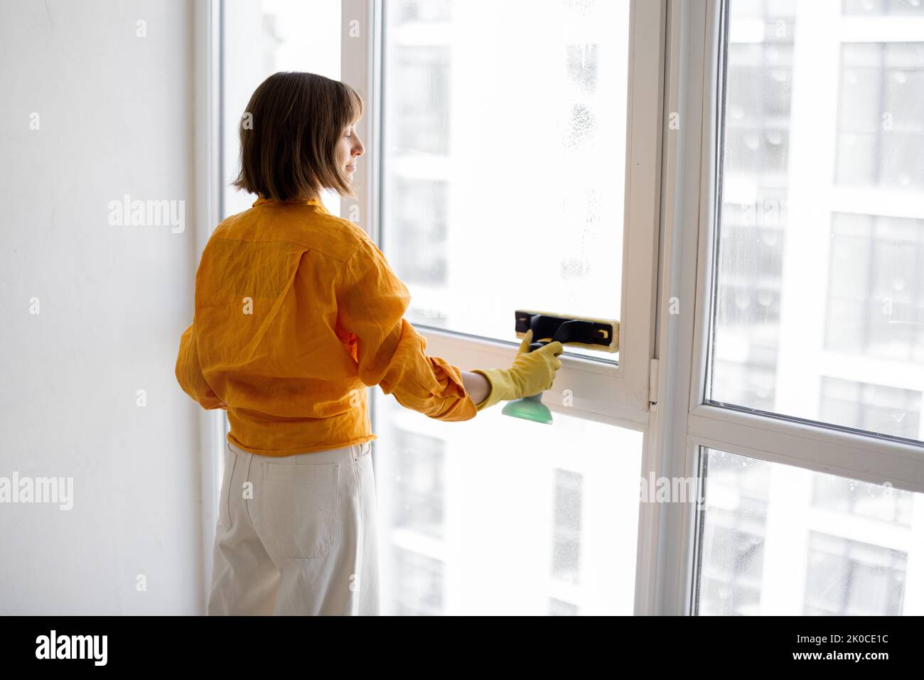 Young housewife or a cleaning company employee washes window with special tool in apartment. Stock Photo