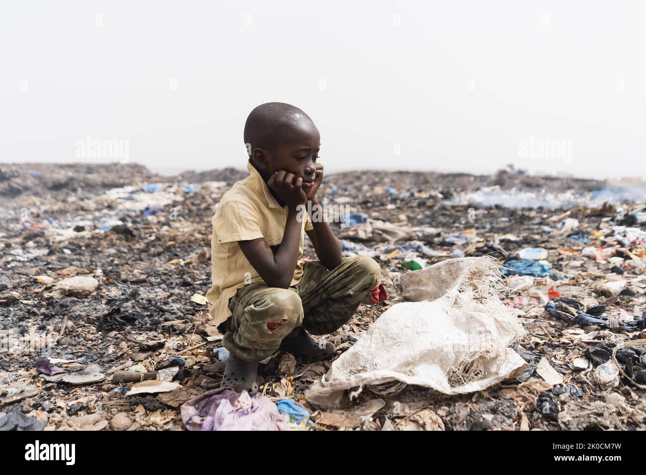 Tired little African boy is resting from the tedious work of collecting garbage at a fuming city garbage dump Stock Photo