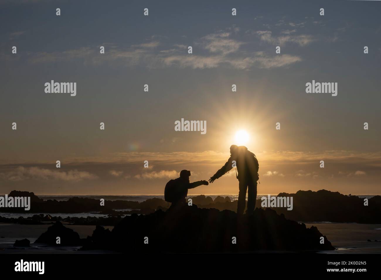 Hiker giving his hands helping partner climb up the rocks at sunrise. Stock Photo