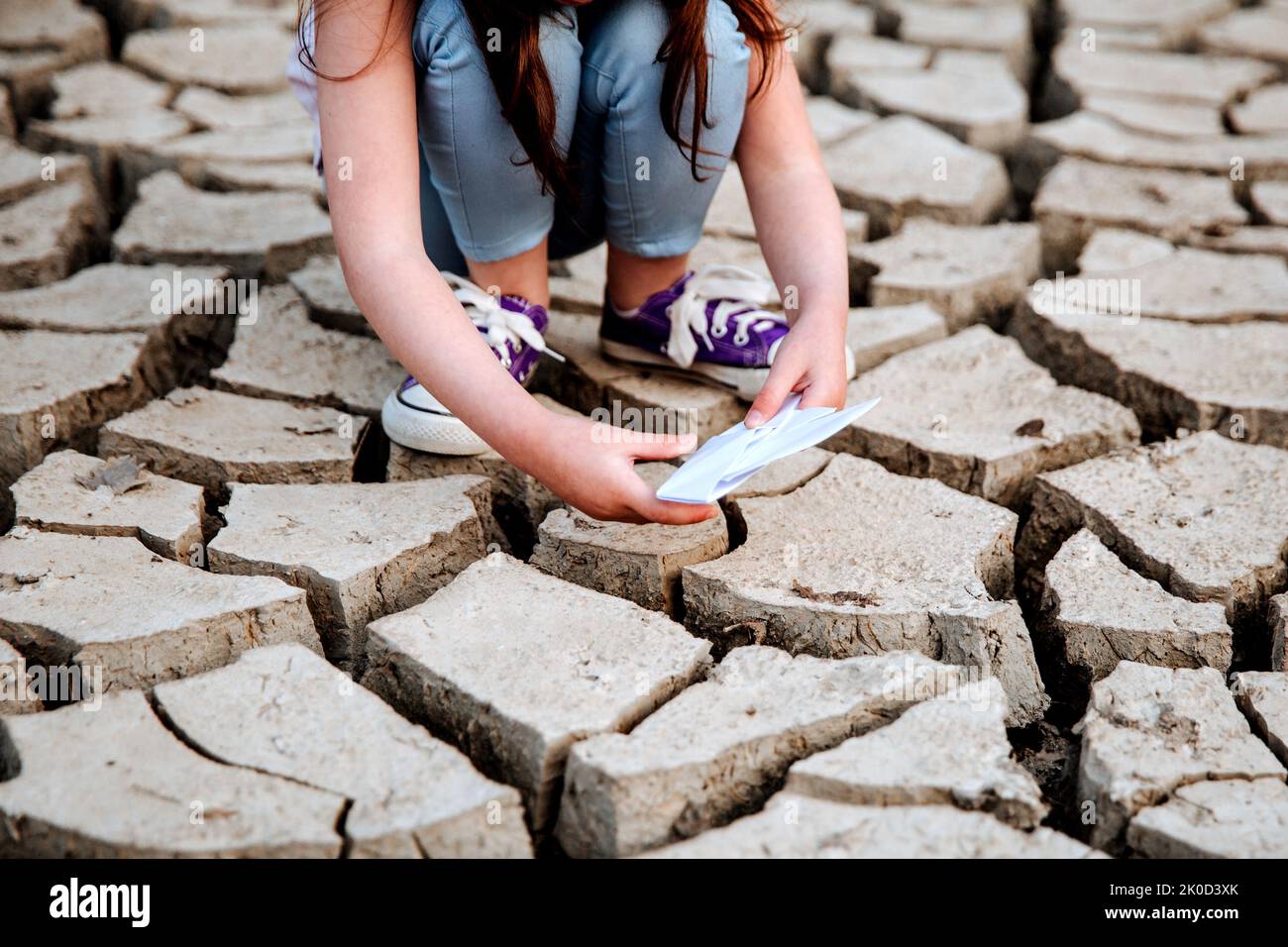The girl lowers the paper boat onto the dry, cracked ground. Water crisis and climate change concept. Global warming Stock Photo