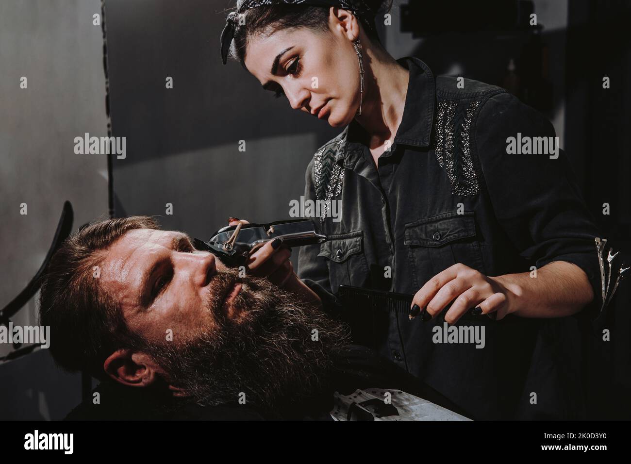 Women professional hairdresser serving client with thick big beard by clipper, handsome brutal stylish young man getting trendy haircut , black barber shop room Stock Photo
