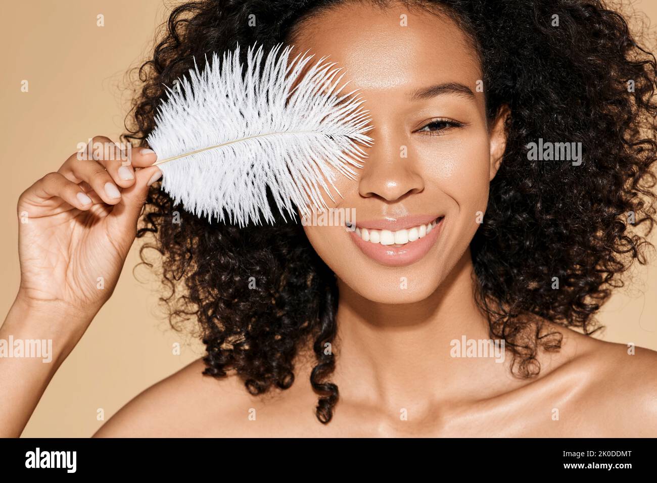 African woman showing how smooth and soft her skin is by touching her face with white feather on beige background. Facial skin care Stock Photo
