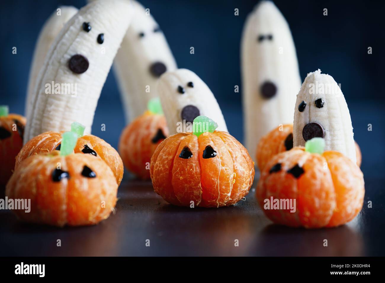 Halloween cute pumpkin orange fruit with scary banana ghosts monsters with chocolate faces behind them. Healthy dessert snack with funny faces. Stock Photo