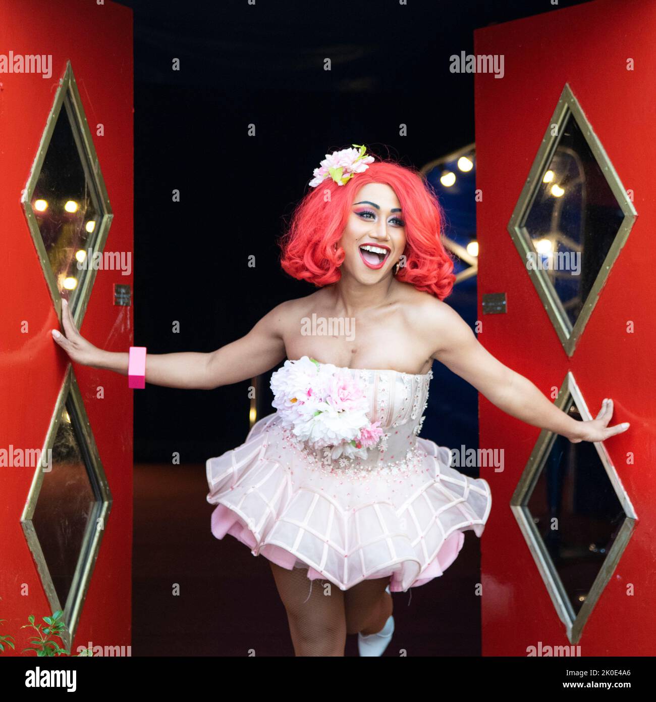 Ladyboy with red wig and pink tutu dress coming out of red doors and smiling, at the ladyboys of Bangkok show Stock Photo