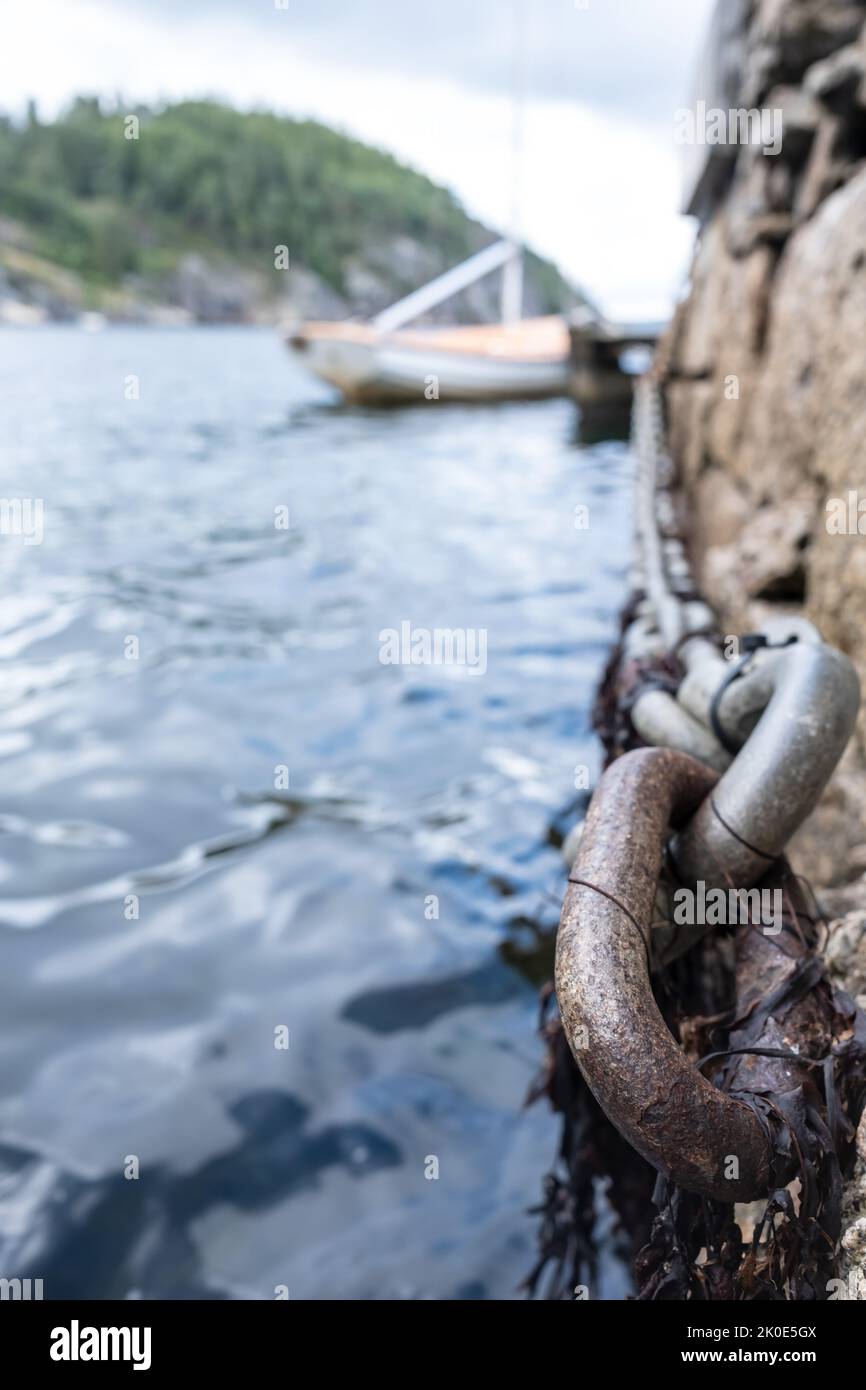 Iron chain on old stone pier, sea waves and moored sailboat and rocky shore out of focus. Bottom view. Stock Photo