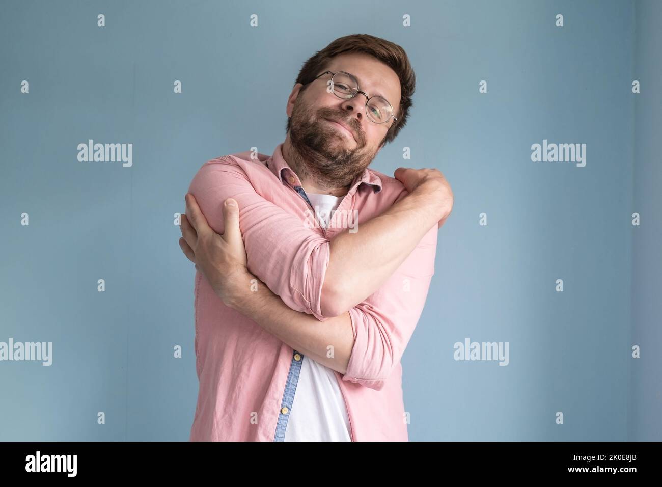 Dreaming about love, a man in glasses and a shirt gently hugs himself, standing on a blue background. Stock Photo