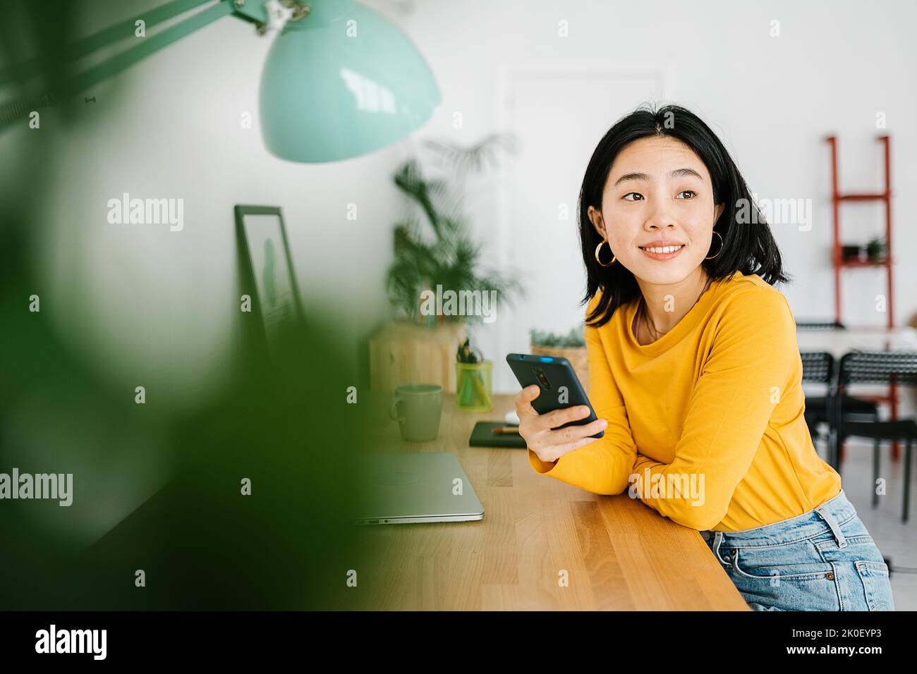 Smiling young asian woman holding mobile phone while working on laptop at home Stock Photo