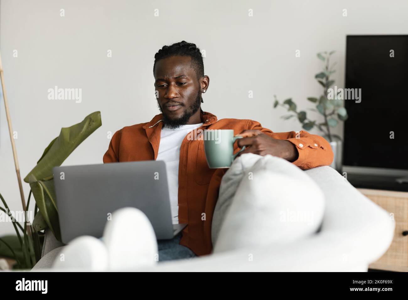 African american man working on laptop, holding cup and enjoying drinking morning coffee, sitting on sofa at home Stock Photo