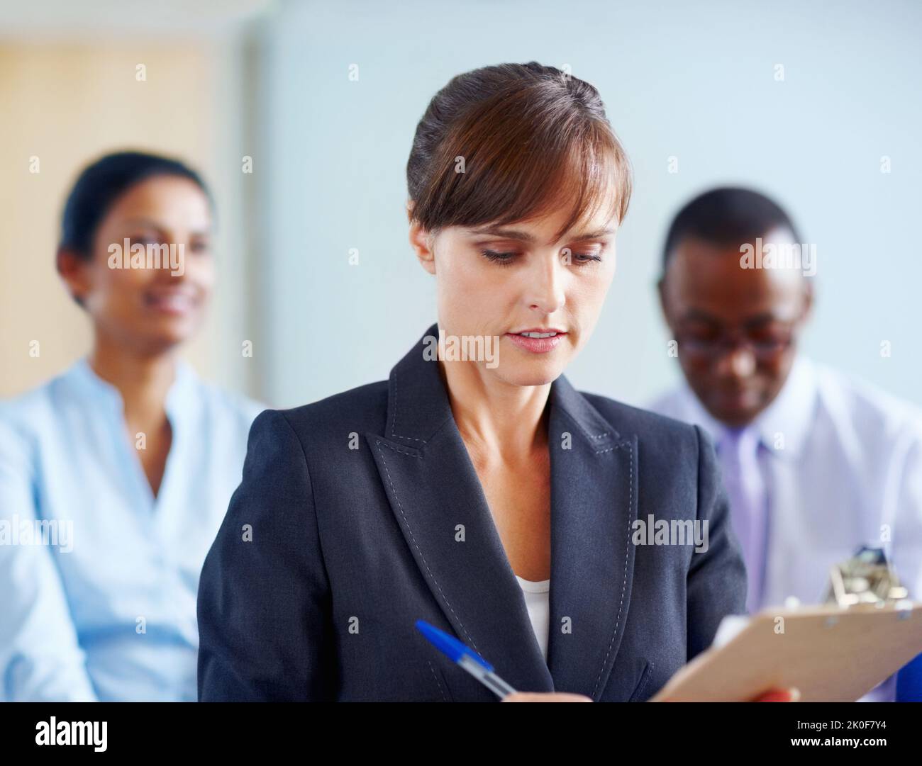 Female executive taking down notes. View of female executive making notes with colleagues in background. Stock Photo