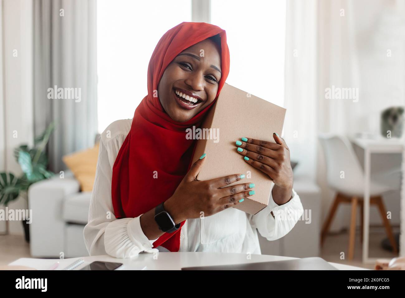 Contented buyer, shopaholism and commerce concept. Happy african american lady in hijab hugging cardboard box Stock Photo