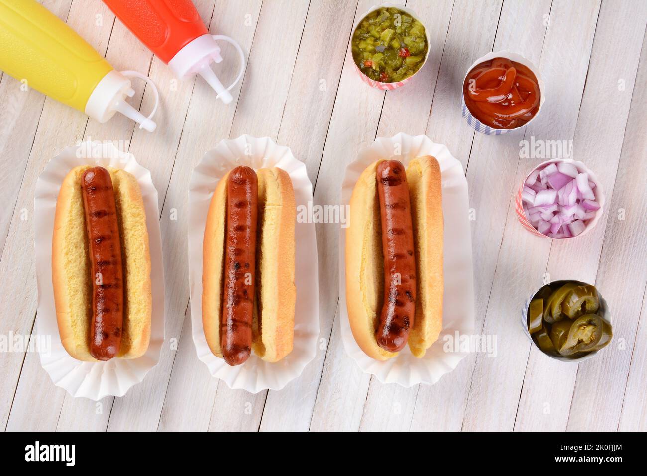 Three grilled hot dogs ready to be garnished on a rustic wood picnic table. High angle view. Stock Photo