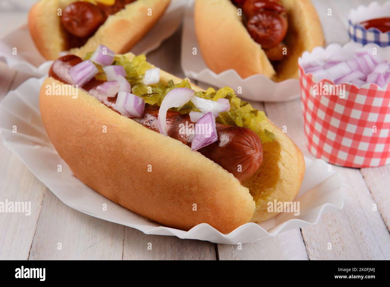Closeup of a hot dog with relish and onions, in the background are two additional franks in buns and cups of condiments. Stock Photo