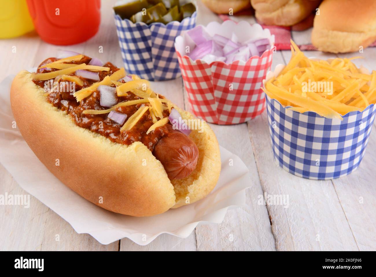 Closeup of a grilled chili dog with cheese and onions on a rustic wood picnic table. More buns and condiments fill the background. Stock Photo