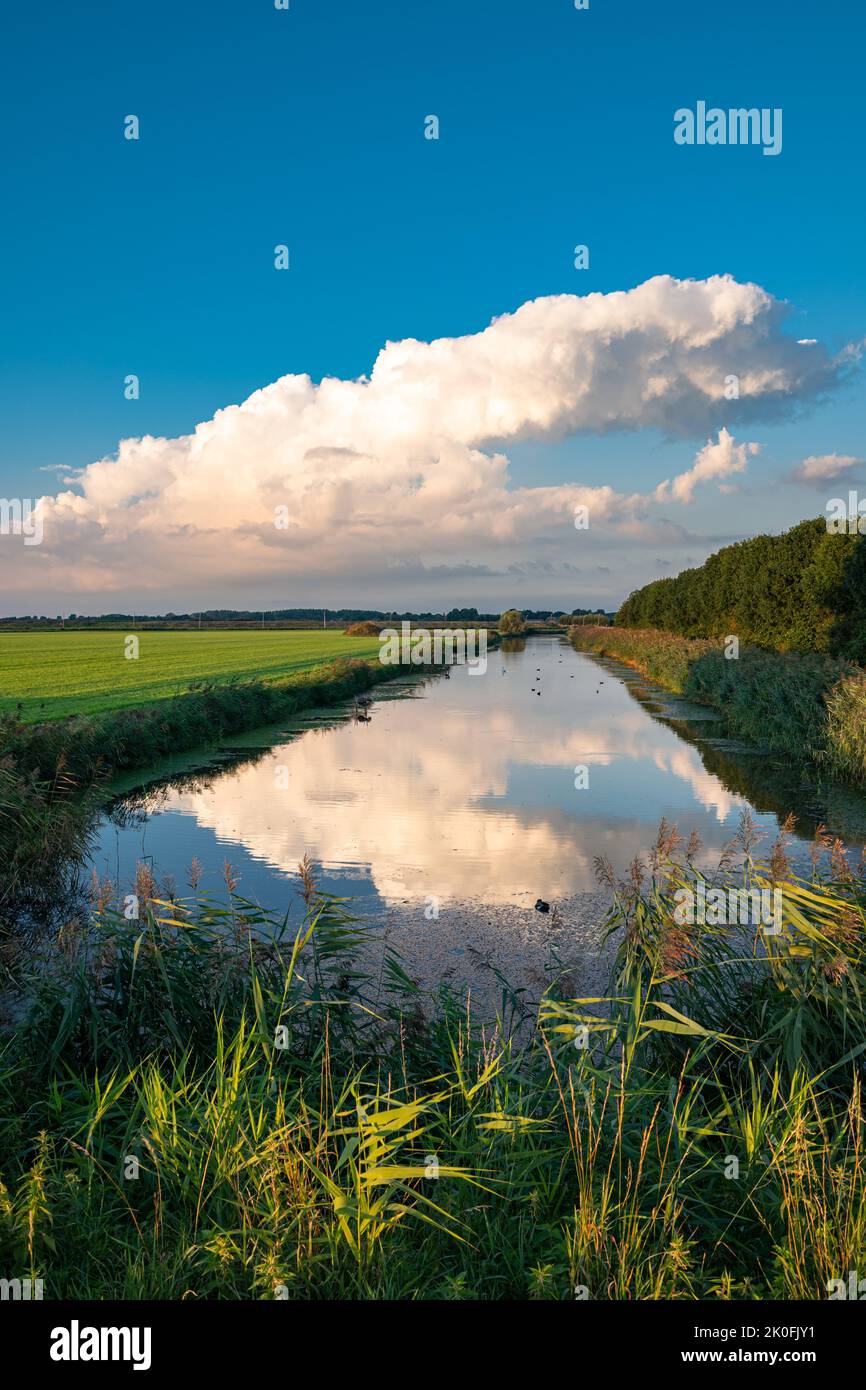 Dissipating storm cloud is reflected in the calm water of a canal on a serene summer evening Stock Photo